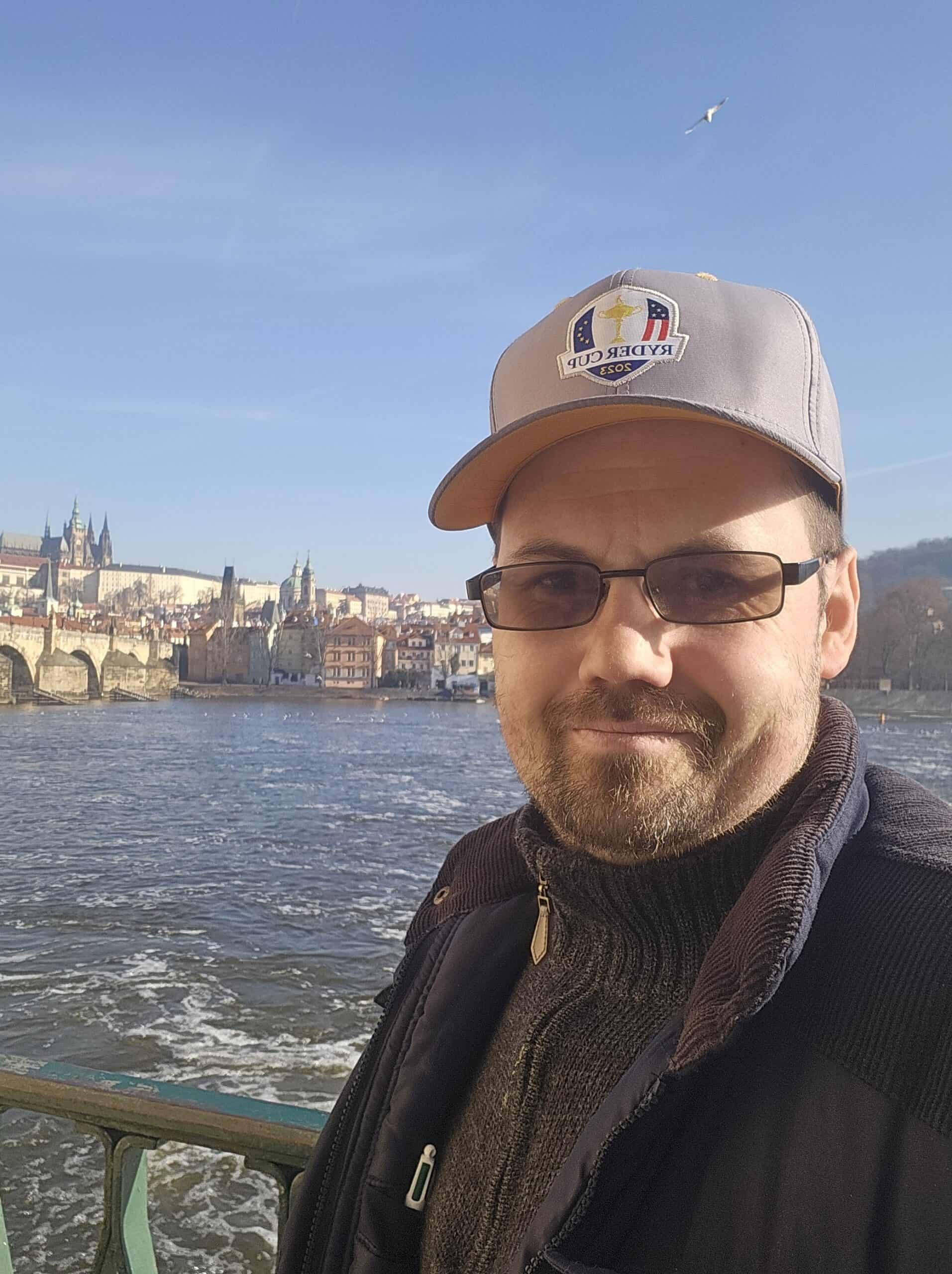 Man wearing golf cap with greenkeeping logo near river in city skyline.