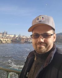 Man wearing golf cap with greenkeeping logo near river in city skyline.